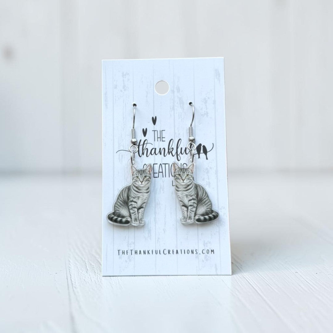 A pair of dangling earrings shaped like sitting tabby cats, displayed on a white branded card that reads 'The thankful creations.' The earrings have silver-toned hooks and gray-brown cat bodies with darker striping.