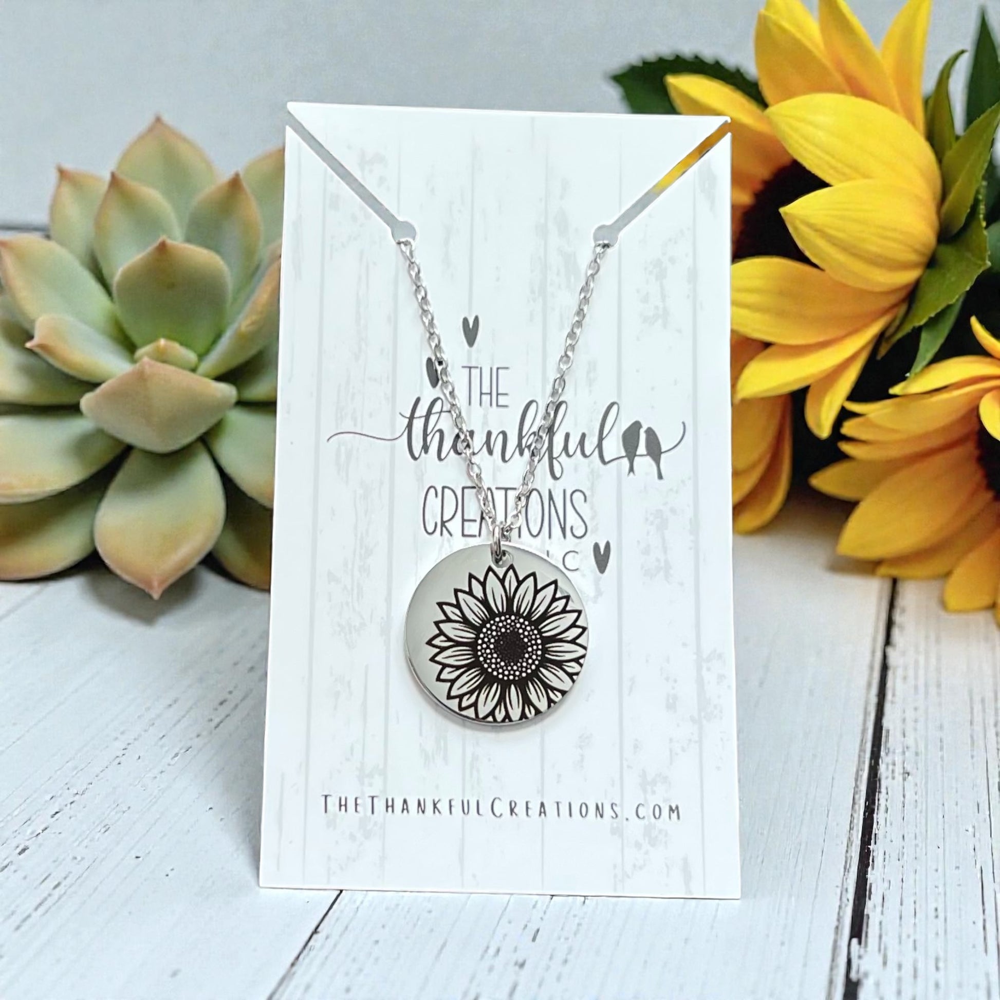 Necklace with sunflower pendant on a card labeled 'The Thankful Creations' with sunflowers and succulents in the background.