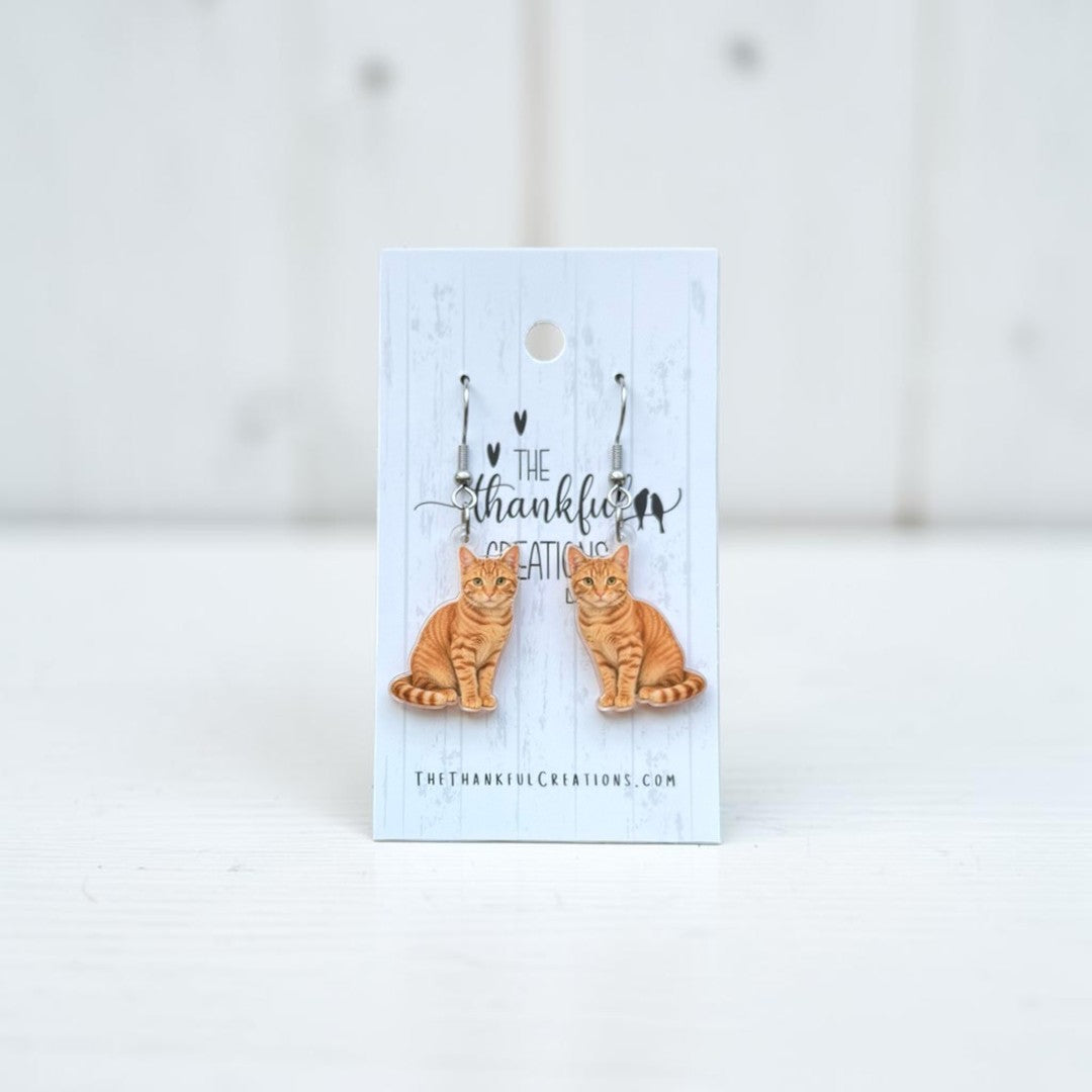 A pair of dangling earrings shaped like orange tabby cats, displayed on a light blue card that reads 'The thankful creation.' The card is photographed against a soft white background.